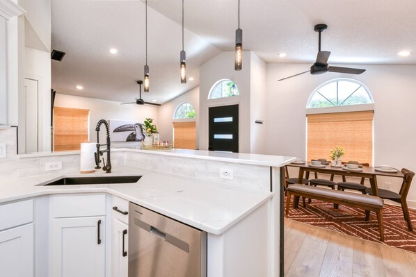 A modern kitchen with white cabinetry, a stainless steel dishwasher, pendant lights, and a dining area with a bench and table.