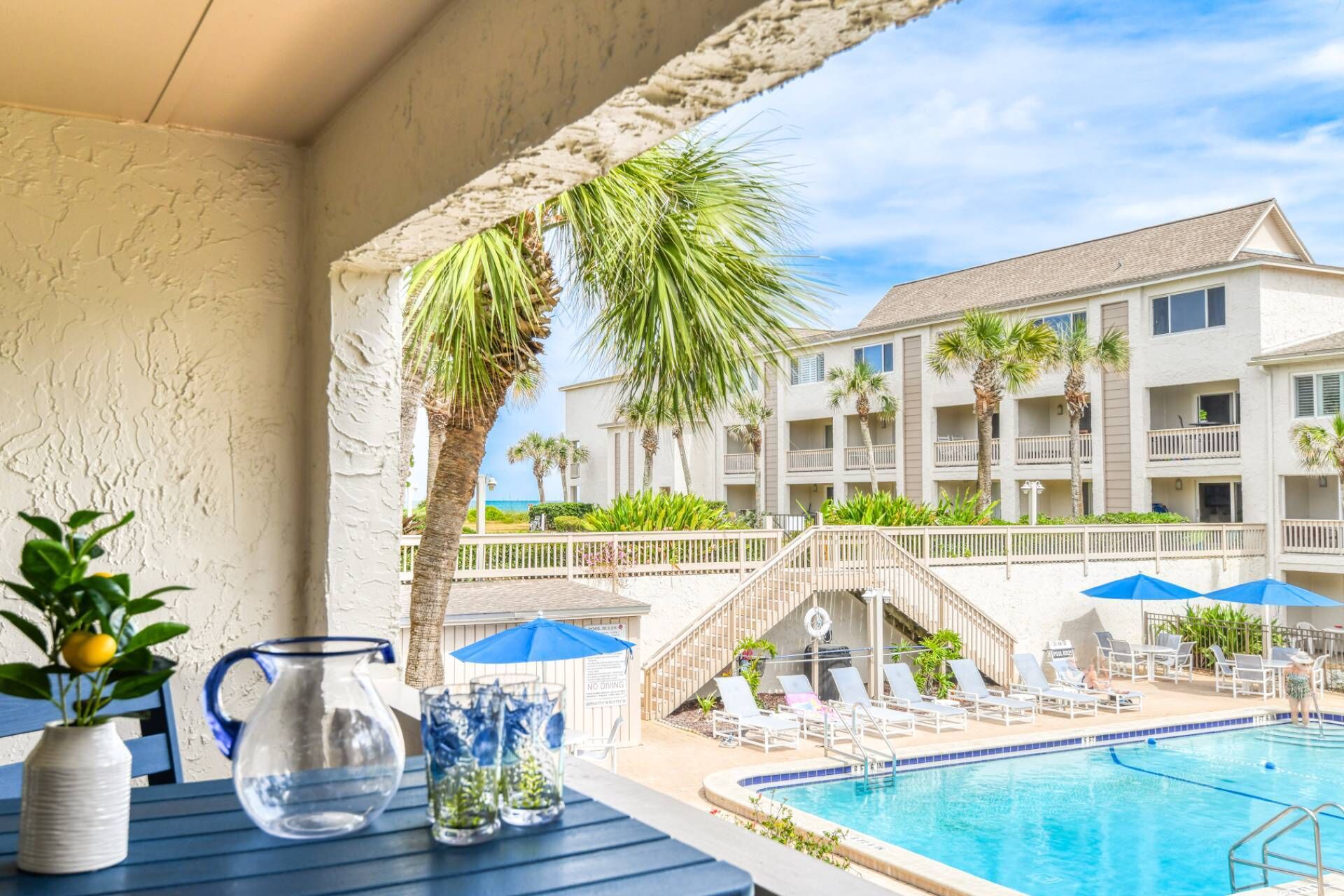 A poolside view with a clear blue pool, lounge chairs, palm trees, and a building with balconies in the background.