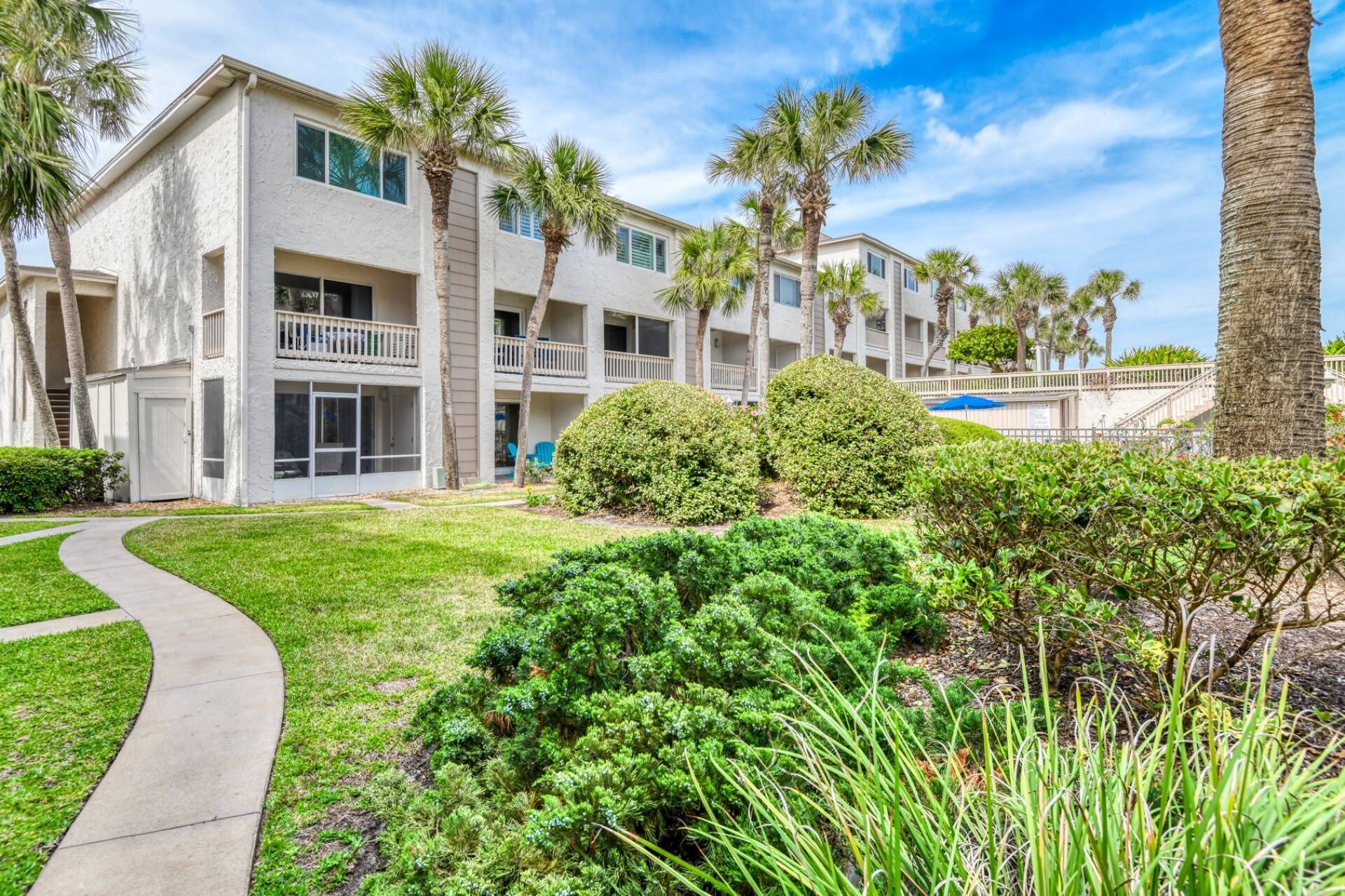 A multi-story building with balconies, a paved walkway, and well-maintained landscaping including palm trees and shrubs.