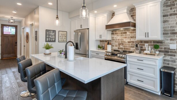 Cocina moderna con armarios blancos, una isla central con asientos, electrodomésticos de acero inoxidable y una pared decorada con ladrillos.