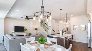 Una cocina moderna con una isla central, un comedor con sillas blancas y una mesa redonda, y una sala de estar con un sofá y una televisión.