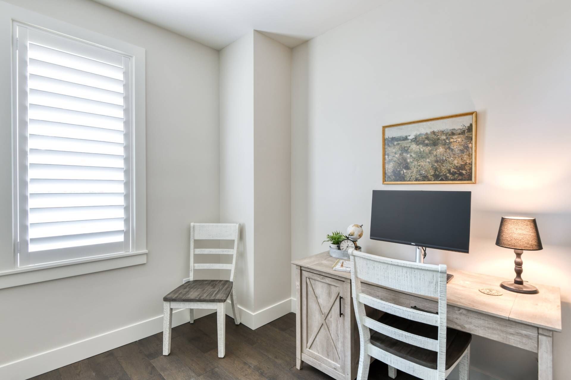 Een hoekappartement met een witte stoel, een bureau met een flatscreen-tv, een lamp en een ingelijst schilderij aan de muur.