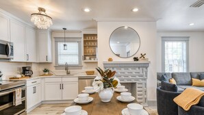 Una cocina moderna con armarios blancos, una mesa de comedor de madera dispuesta con vajilla blanca, una araña y una chimenea.