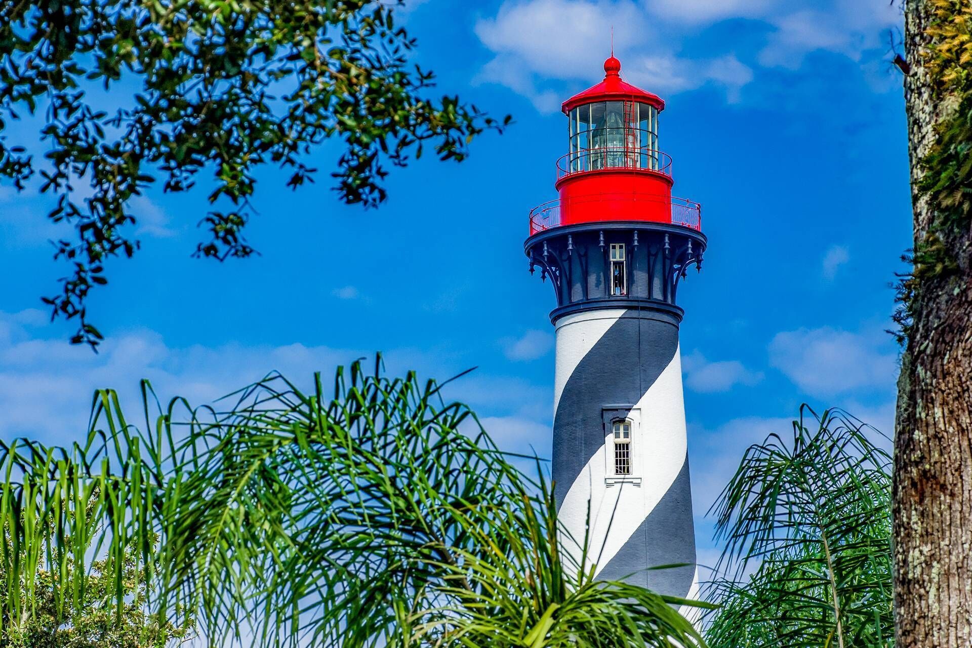 A lighthouse with a red top and white and black striped body, surrounded by greenery.