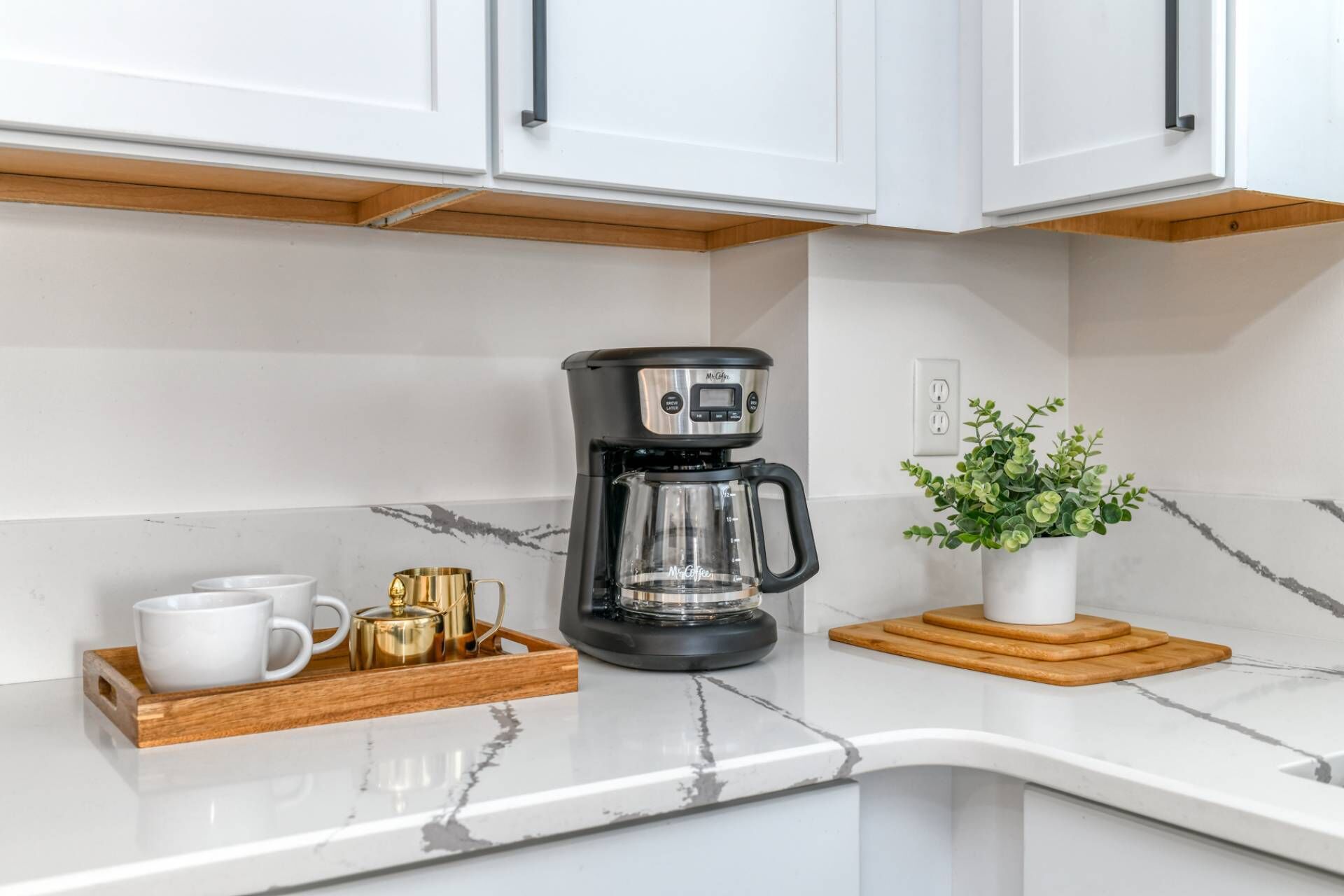 Una cocina con armarios blancos, una cafetera, una planta en maceta y una bandeja con tazas y una tetera.