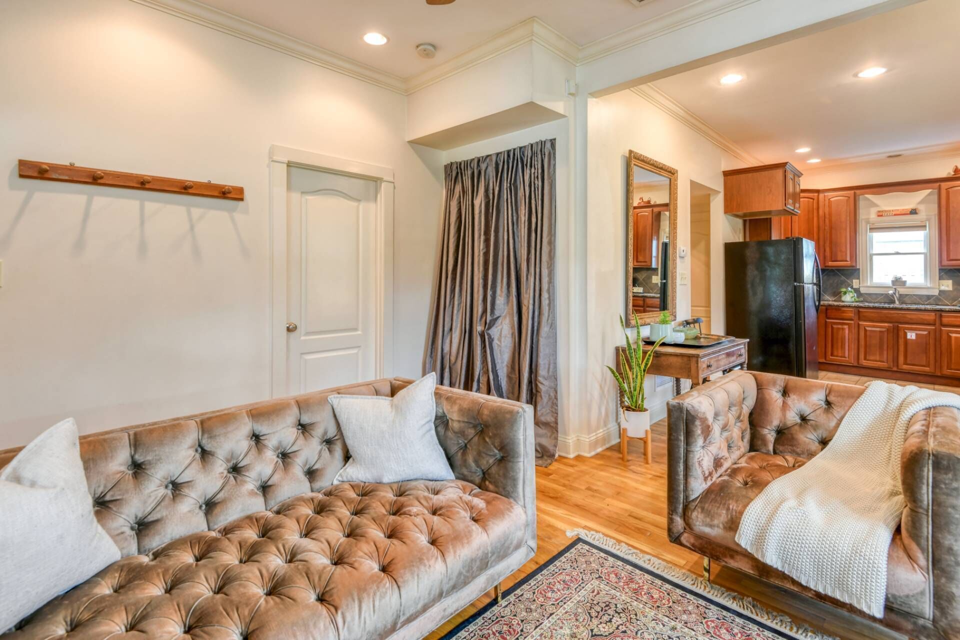 A spacious living room with two tufted sofas, a large window with curtains, and a kitchen area with wooden cabinets and a black refrigerator.