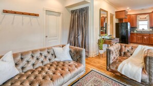 A spacious living room with two tufted sofas, a large window with curtains, and a kitchen area with wooden cabinets and a black refrigerator. - Little Blue Cottage (St. Augustine)