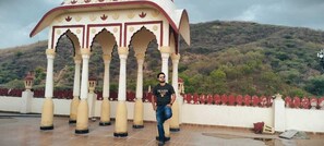 A person standing in front of a traditional structure with arches and columns, set against a backdrop of hills and a cloudy sky.