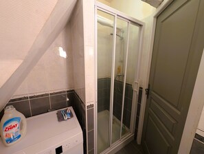 Une salle de bain avec un espace douche, un lave-linge blanc et une bouteille de lessive.