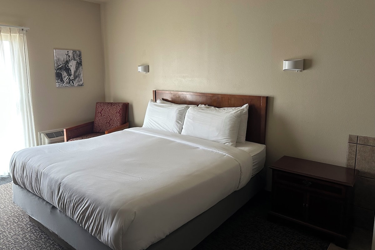 A hotel room with a large bed, a nightstand, a chair, and a wall-mounted lamp.