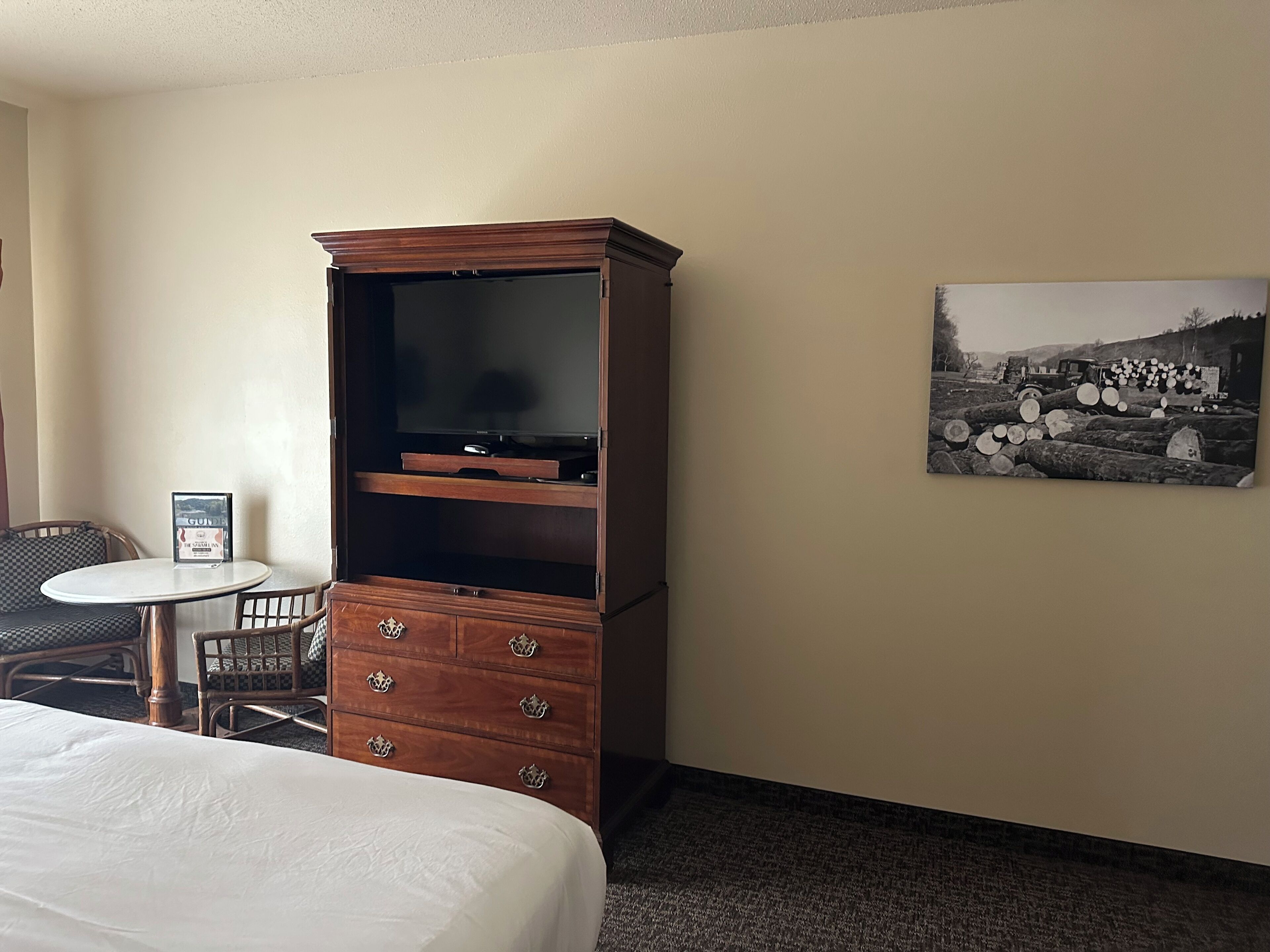 A hotel room with a bed, a wooden cabinet with drawers, a small table with a chair, and a framed picture on the wall.