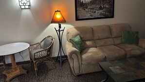 A living room with a beige sofa, a small round table, a wicker chair, a floor lamp, and a framed photograph on the wall. - The Sawmill Inn (Houston)