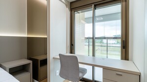 A modern bedroom with a built-in desk, a chair, a bed, and a large window offering a view of the outdoors. - Apart w/ 2 Suites in Jurere Internacional Vvc0202 (Florianópolis)