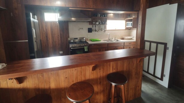 A kitchen with wooden cabinets, a stainless steel stove, a microwave, and a sink. There are two stools at the counter. - Monteverde Monkey Hostel (Monteverde)