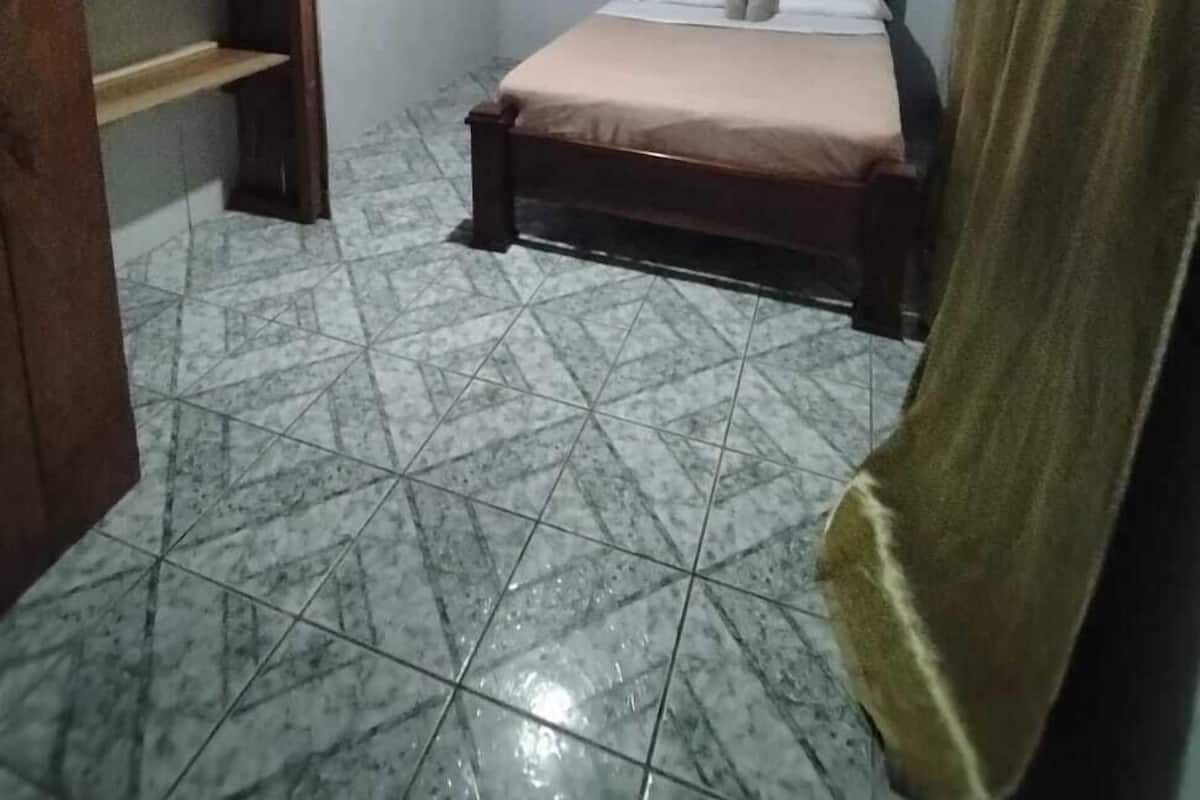 Una habitación pequeña, con poca luz, que tiene una cama individual, una estantería de madera y una cortina.