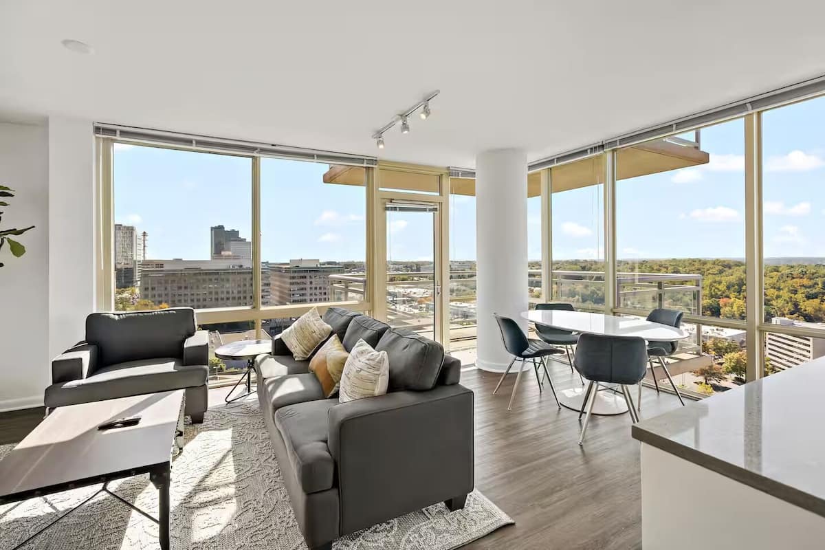 A modern living room with a large sofa, a dining table, and chairs. There are large windows offering a city view.