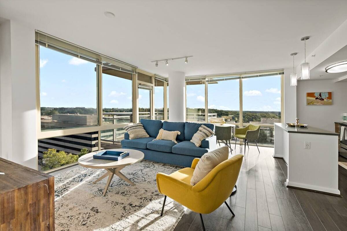 A spacious living area with a blue sofa, yellow armchair, and a coffee table. Large windows offer a view of the cityscape.