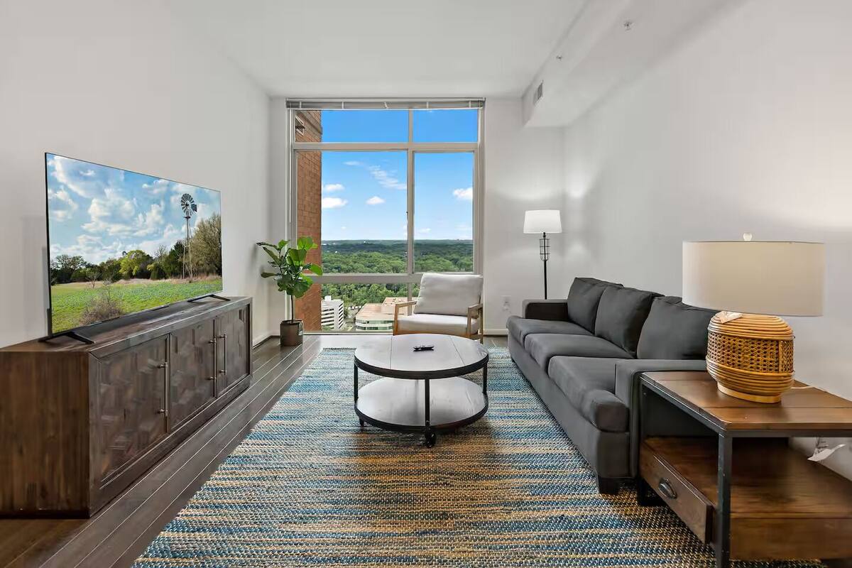 A modern living room with a large TV, a grey sofa, a coffee table, and a view of a green landscape through large windows.