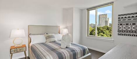 A modern bedroom with a large bed, two bedside tables, a window with a city view, and a patterned wall decoration.