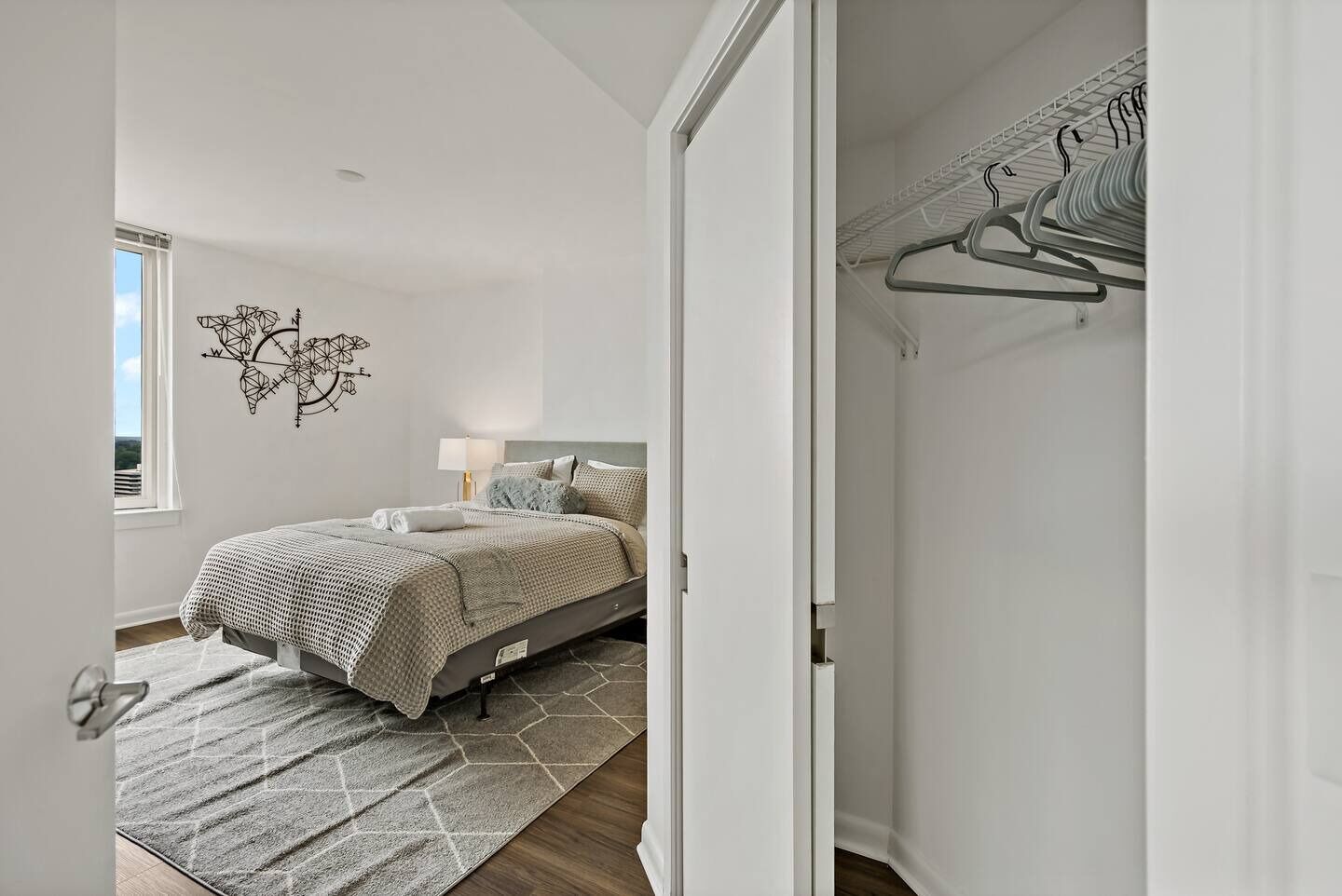 A bedroom with a bed, a geometric wall decoration, a bedside lamp, and a view of a closet with hanging clothes.