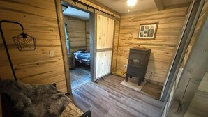 A wooden cabin interior with a bed, a small heater, a chair, and a sliding door leading to another room.