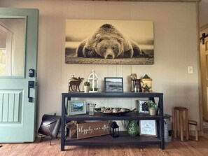 Una habitación con una consola de madera que exhibe varios objetos, un cuadro colgado con un oso y una puerta con marco azul claro.