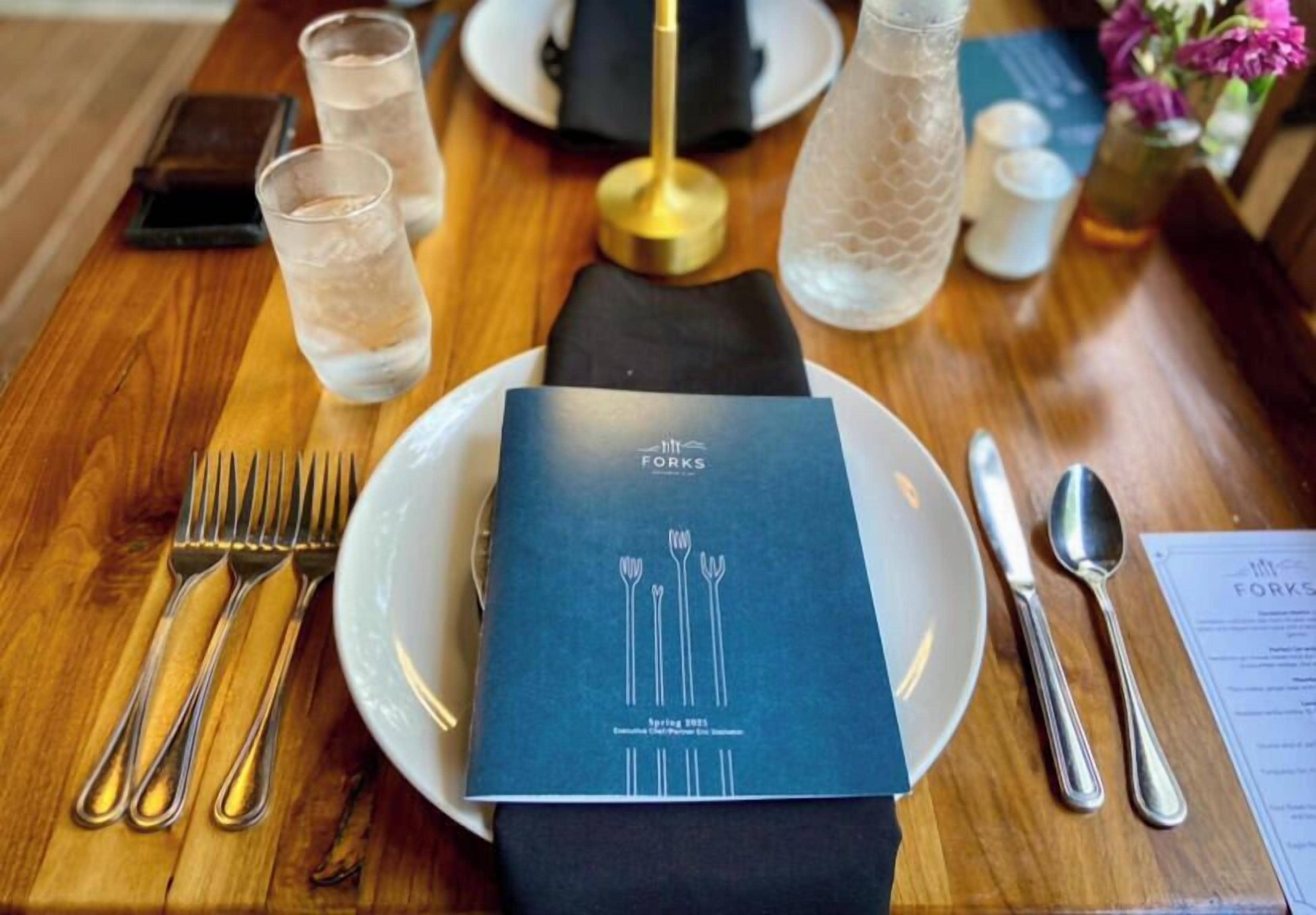 A neatly set dining table with a menu, glasses of water, cutlery, and a decorative bottle.