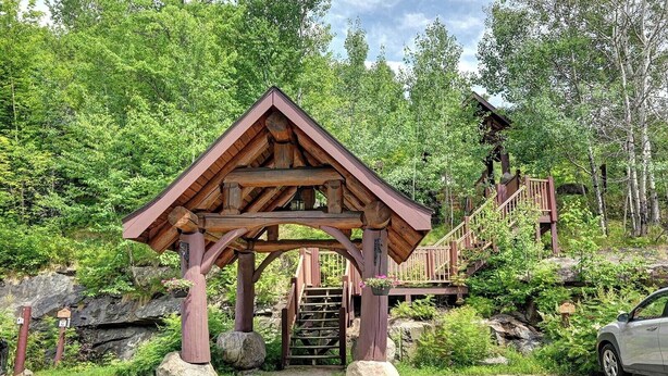 A wooden gazebo with a triangular roof, surrounded by lush greenery and a wooden staircase leading to a higher platform. - 120 Algonquin Unit 7 (Mont-Tremblant)