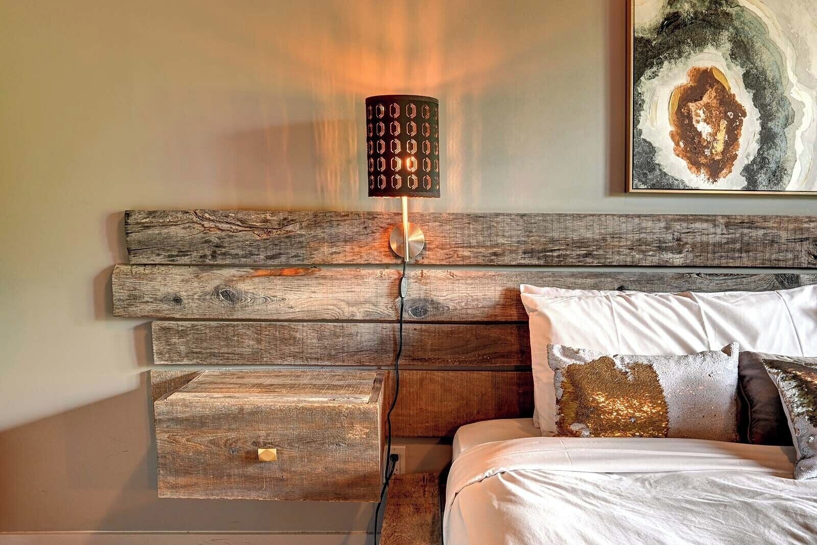 A bedroom with a wooden headboard, a bedside table, a lamp, and a bed with pillows.