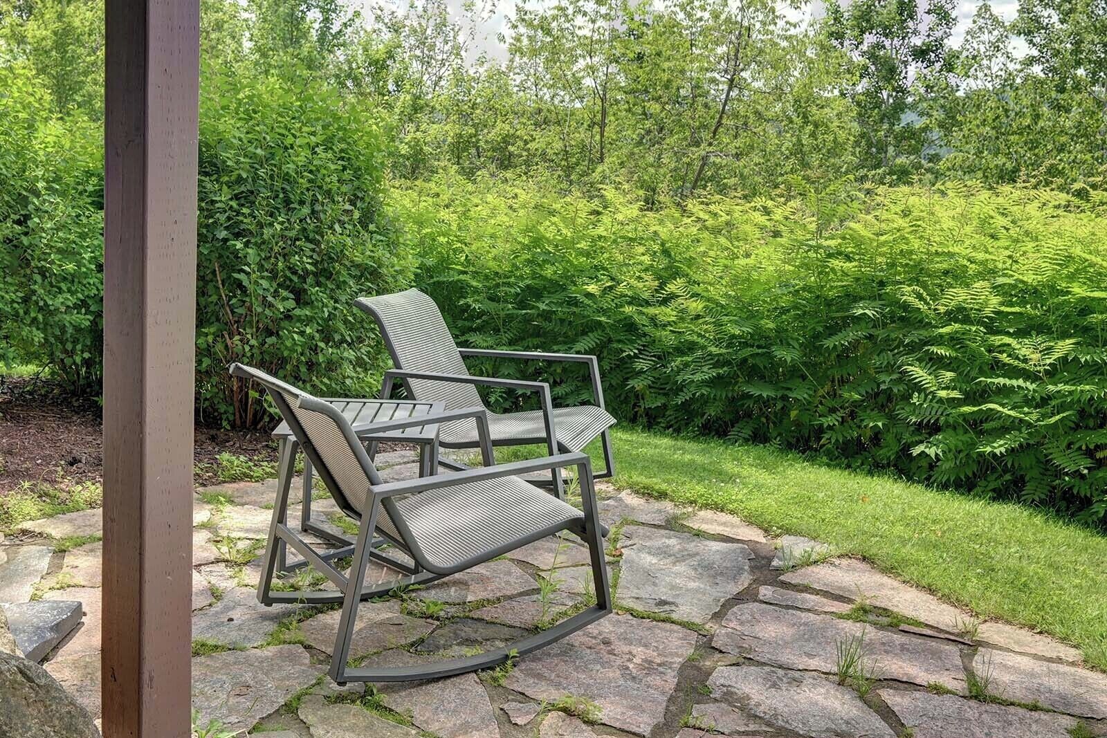 Een terras met twee metalen stoelen en een tafel, omringd door een stenen pad en weelderige beplanting.