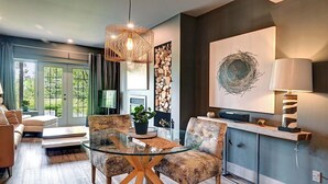 A modern living room with a glass dining table, two upholstered chairs, a hanging light fixture, a fireplace, and a large abstract painting on the wall. - 120 Algonquin Unit 7 (Mont-Tremblant)