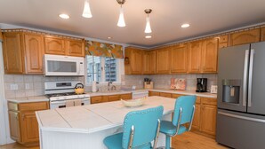 A kitchen with wooden cabinets, a white island with bar stools, stainless steel appliances, and pendant lighting. - Edgartown Escape – Walk to Beach & Downtown (Edgartown)