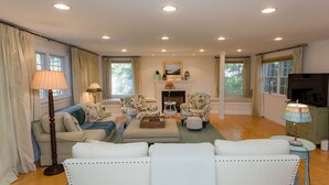 A spacious living room with a fireplace, two floral armchairs, a sofa, and a television. - Edgartown Escape – Walk to Beach & Downtown (Edgartown)