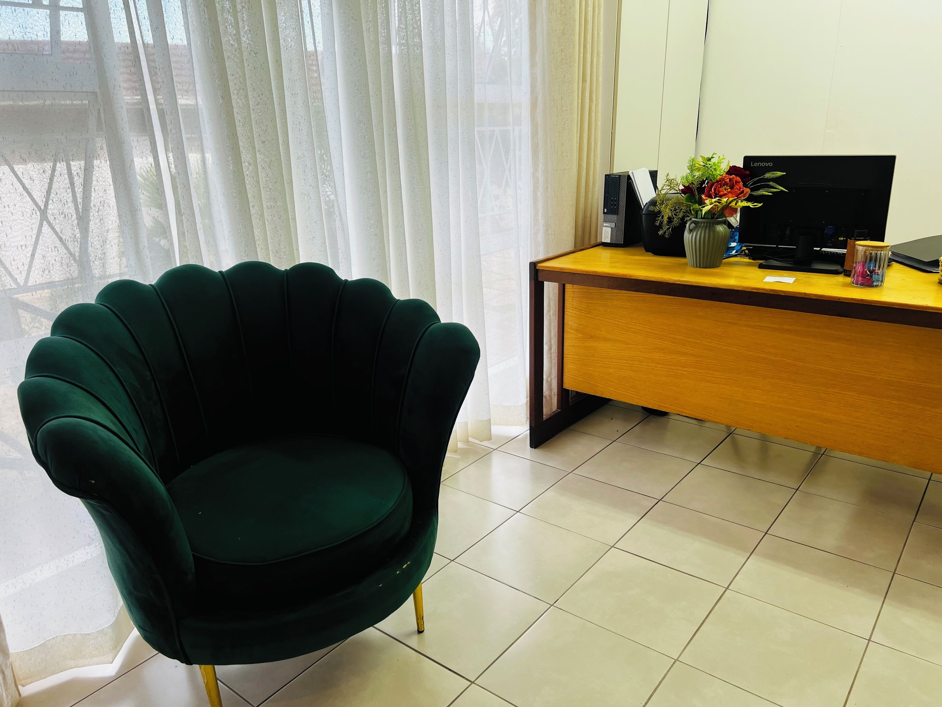 A room with a green armchair, a wooden desk with a laptop, and a vase with flowers.