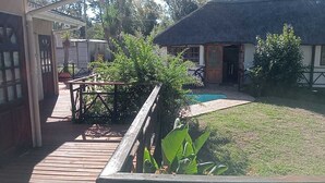 Una terraza de madera que conduce a un edificio con techo de paja, una pequeña piscina y un jardín cubierto de césped con plantas en macetas.
