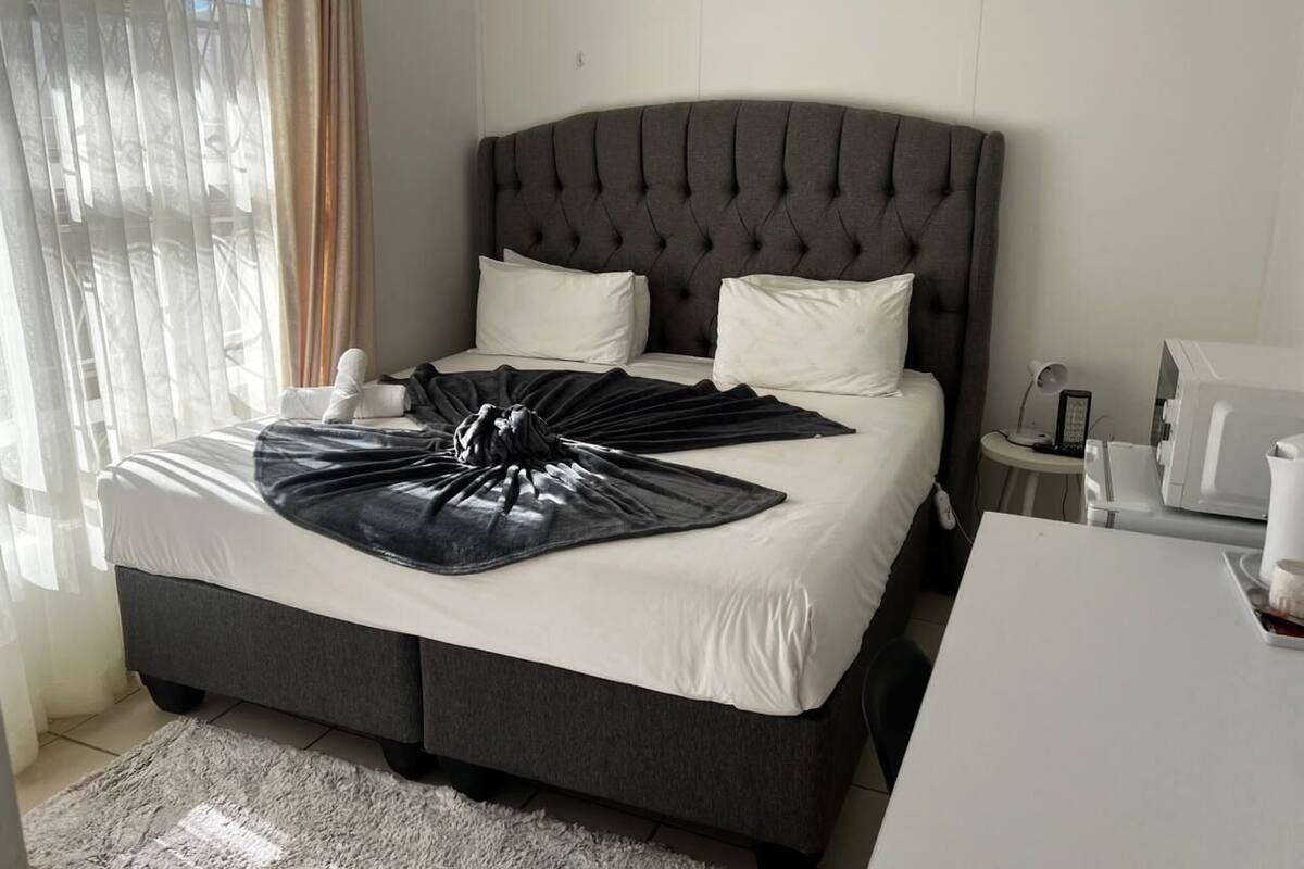 Een netjes opgemaakt bed met een donker hoofdbord, witte kussens en een zwarte deken. Er staat een klein nachtkastje met een lamp en een witte muur.