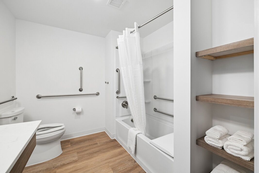 Baño con inodoro, lavamanos, bañera y ducha con barras de apoyo. Hay repisas de madera con toallas.