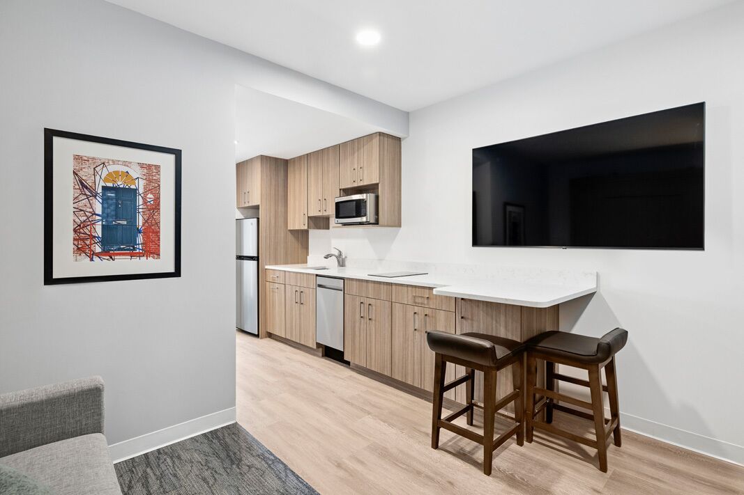 Una cocina moderna con armarios de madera, microondas y encimera blanca. Hay dos taburetes de bar y una foto enmarcada en la pared.