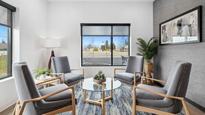 A modern room with a round glass table, four grey chairs, a large window with a view, a framed picture on the wall, and a potted plant.