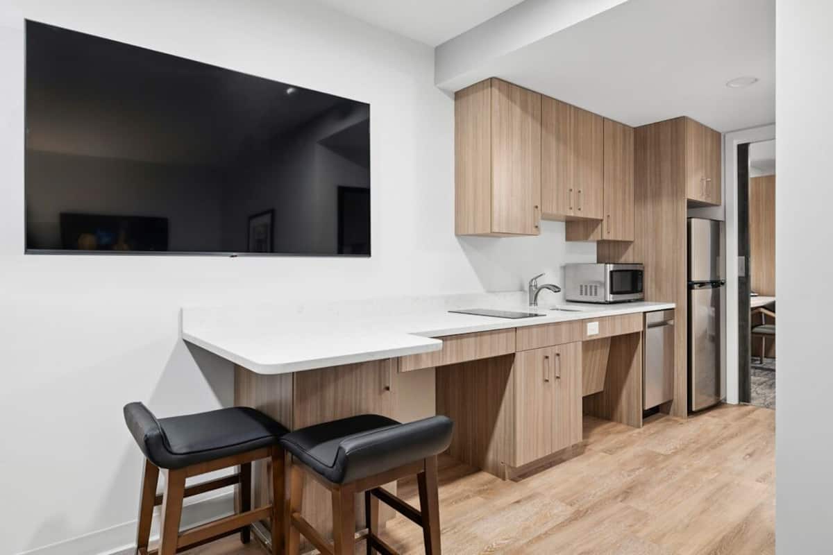 Una cocina moderna con cubierta de encimera blanca, armarios de madera, microondas y refrigerador. Hay dos taburetes negros en la barra.