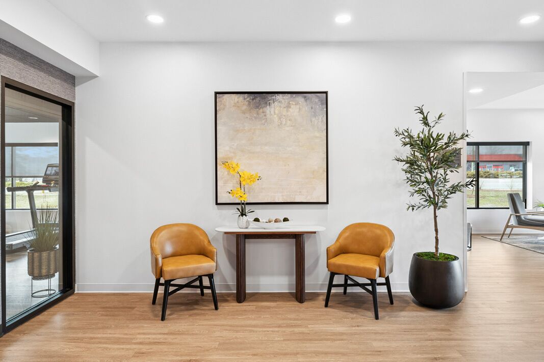 A modern room with a large window, a table with two chairs, a potted plant, and a framed abstract painting on the wall.