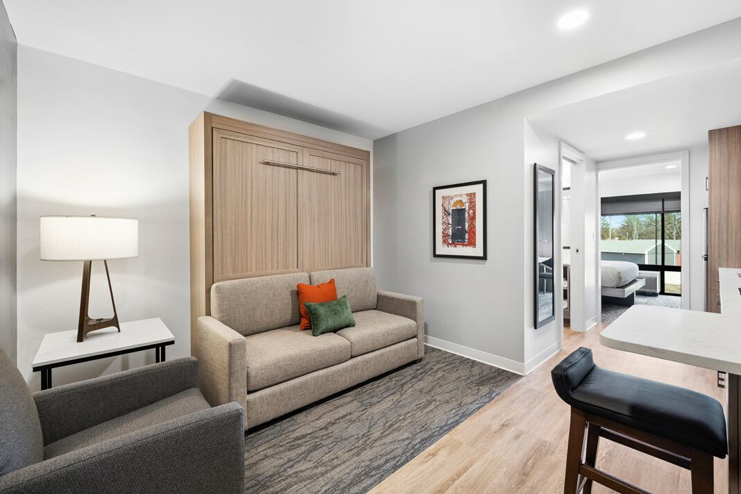 A modern living room with a sofa, a table, and a bench. There is a lamp on a side table, a framed picture on the wall, and a wardrobe.