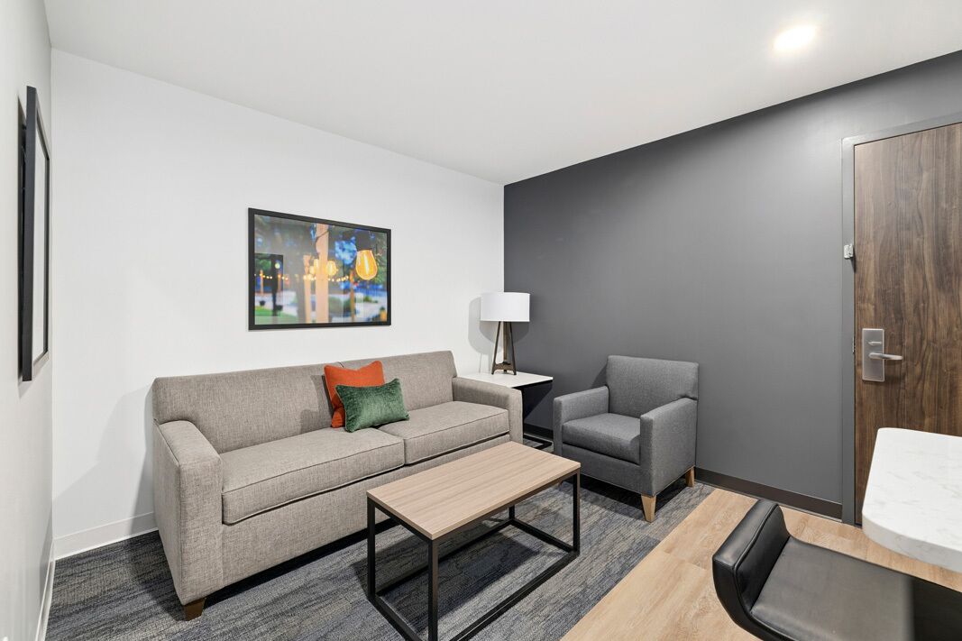 A modern hotel room with a grey sofa, a small table, a grey armchair, a lamp, and a wooden door.