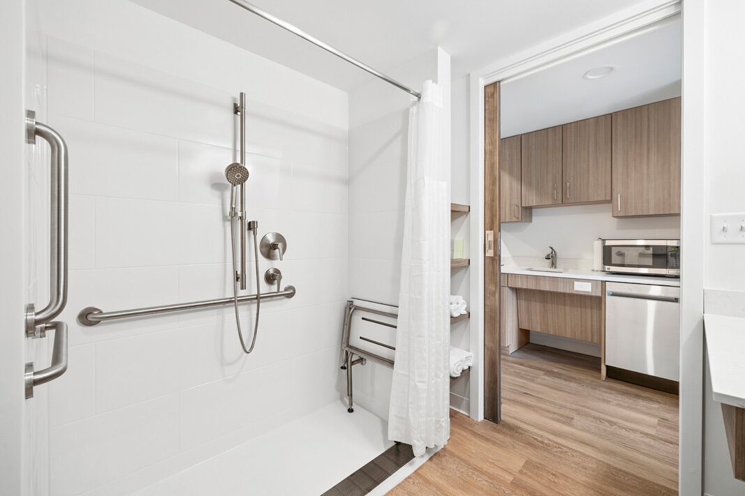 Uma casa de banho moderna com chuveiro, sanita e zona de cozinha visível através de uma porta aberta.