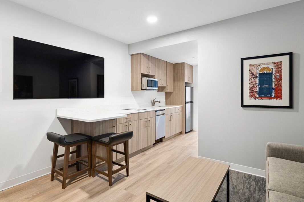 Una cucina moderna con un piano di lavoro bianco, pensili in legno, un forno a microonde e un frigorifero. Sono presenti due sgabelli da bar e un quadro appeso al muro.