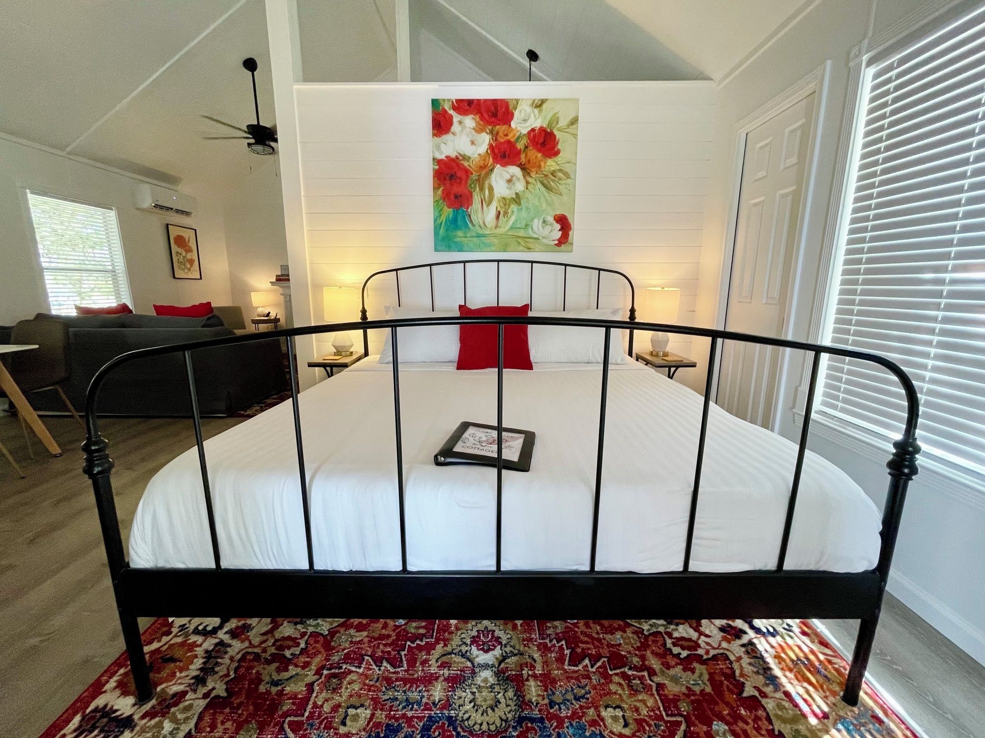 A bedroom with a black metal bed frame, white bedding, a red pillow, a bedside table with a lamp, and a large floral painting on the wall.