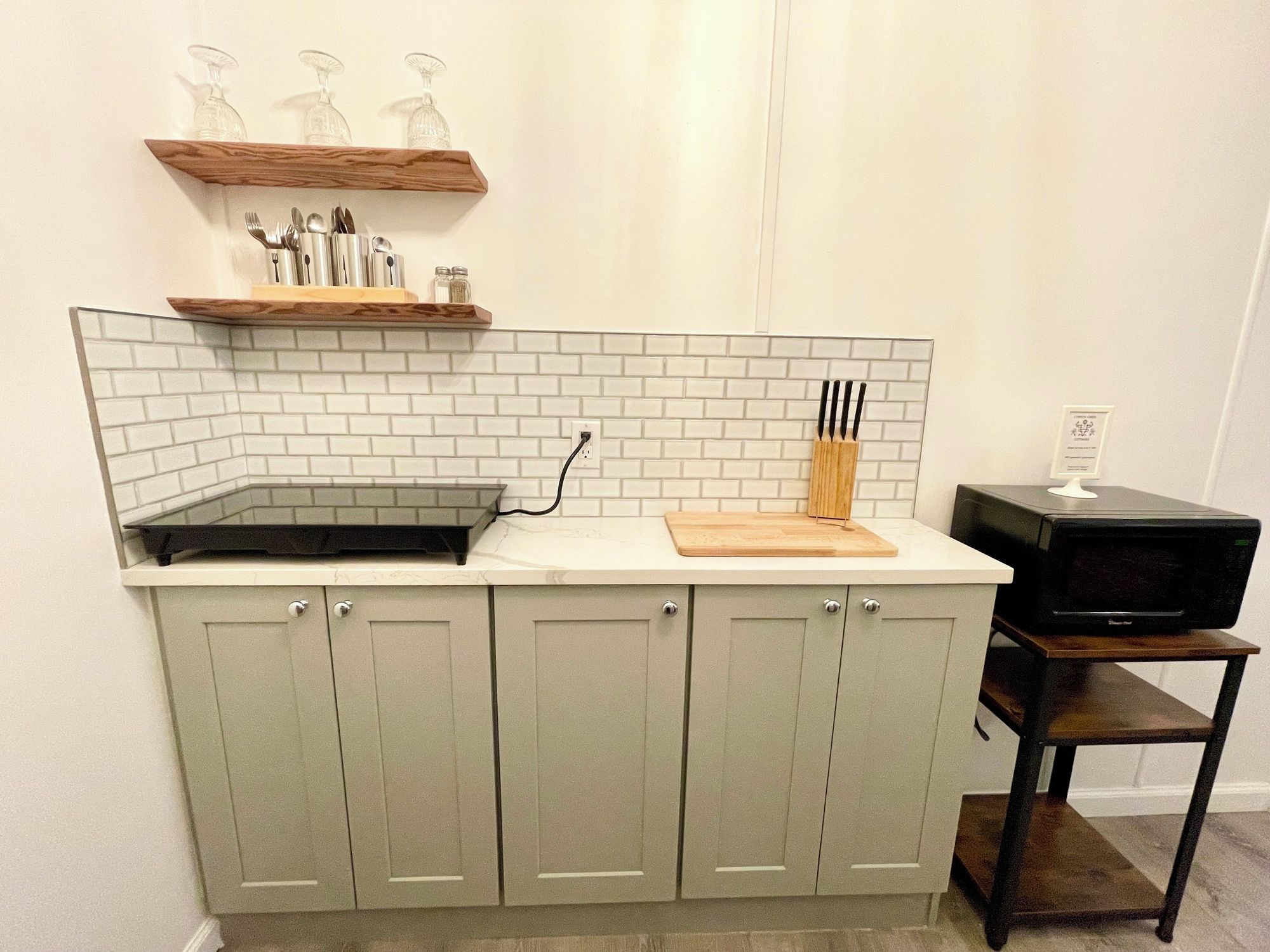 A kitchen with a white tiled backsplash, a wooden shelf above the countertop, and a microwave on a wooden stand.