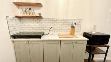 A kitchen with a white tiled backsplash, a wooden shelf above the countertop, and a microwave on a wooden stand.