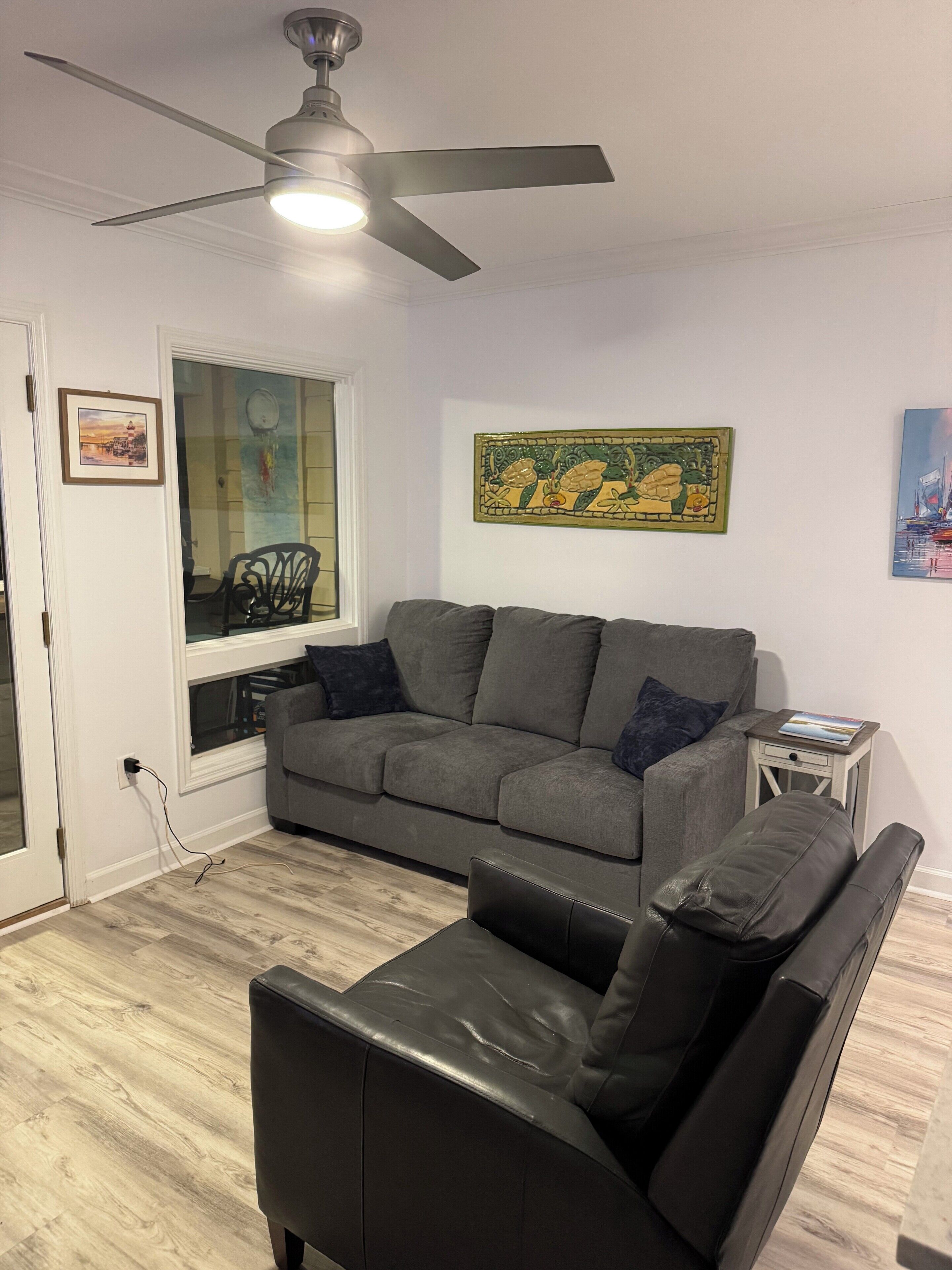 A living room with a grey sofa, a black leather chair, a ceiling fan, and a window with a view of a cityscape.
