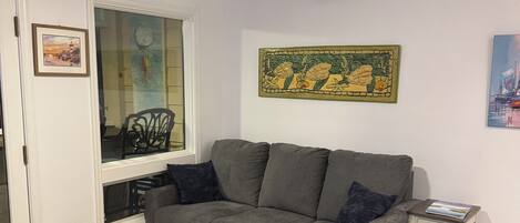 A living room with a grey sofa, a black leather chair, a ceiling fan, and a window with a view of a cityscape.